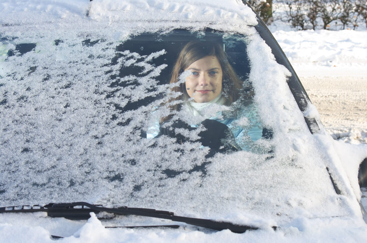 A young woman sitting behind the steering wheel of her car and looking out through her windshield that has only been partly cleared of ice.